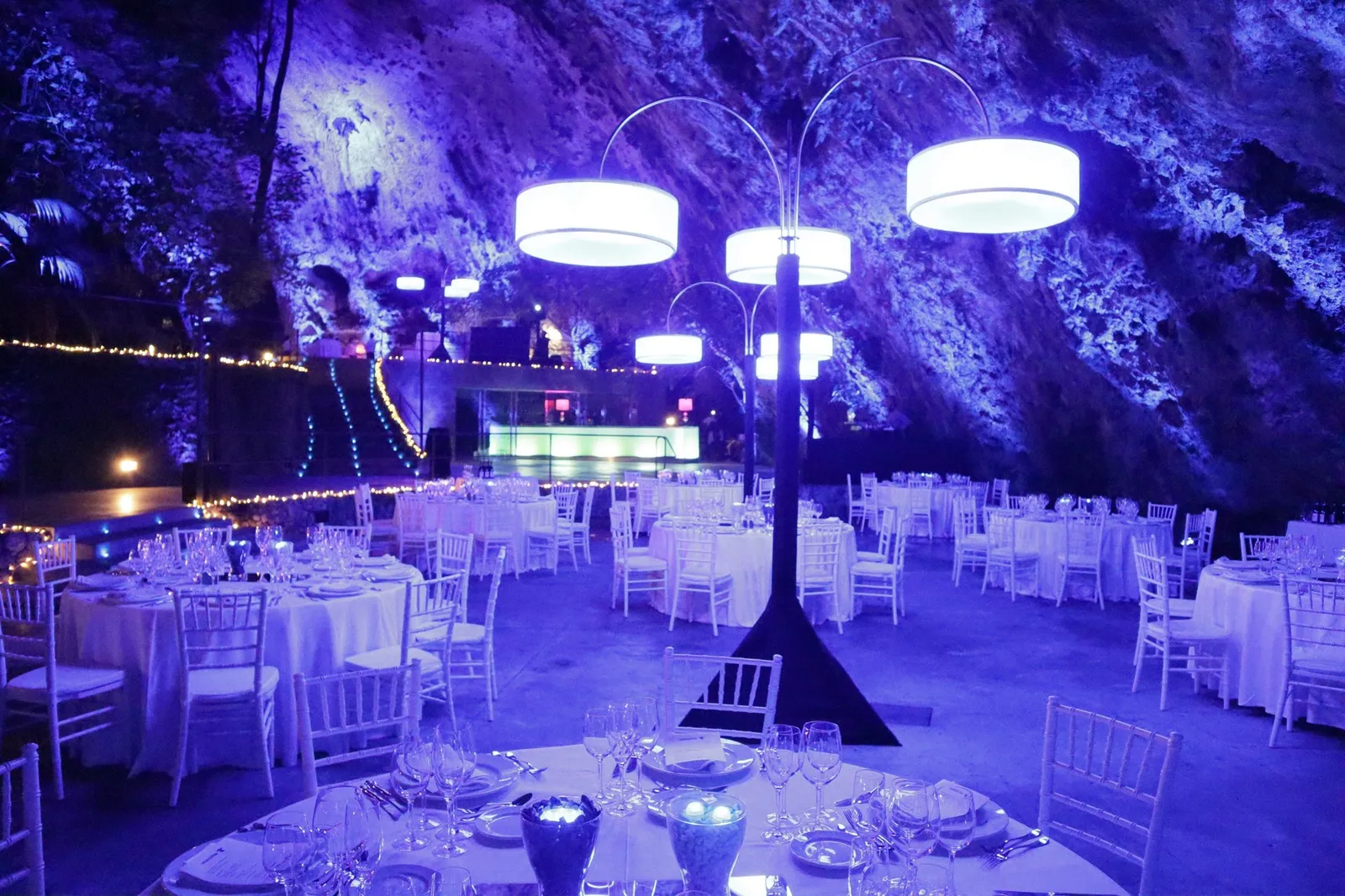 Banquete al pie de una pared rocosa iluminada en azul, con grandes pantallas cilíndricas suspendidas sobre las mesas